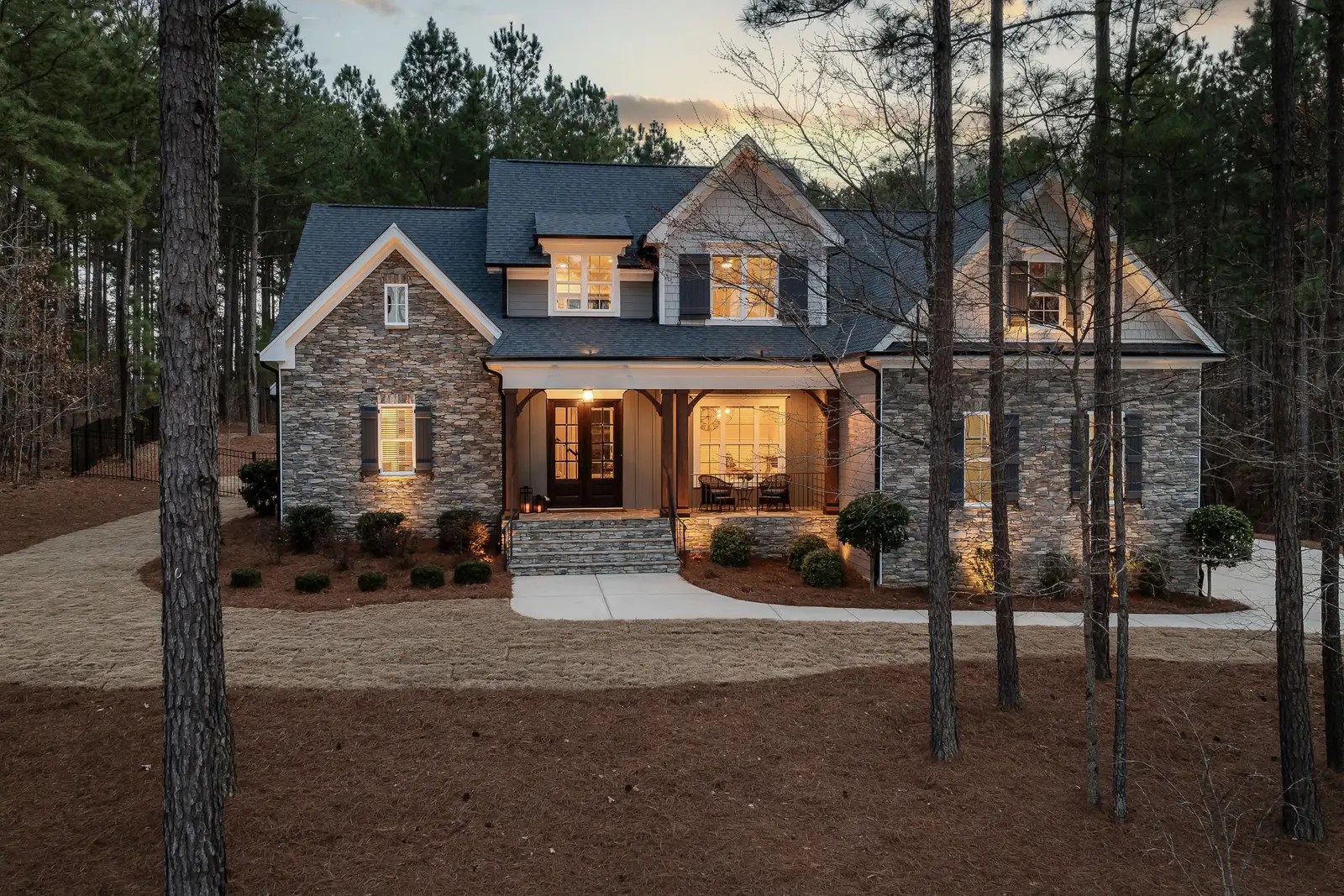 A stone and timber house among tall pines at dusk with warm light in every window